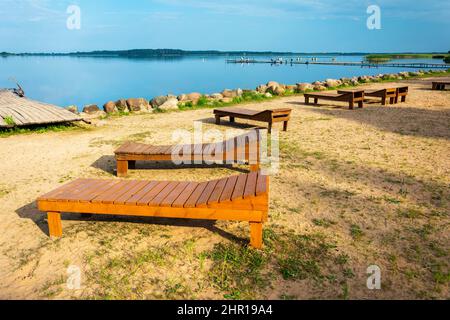 Eine Reihe von hölzernen Sonnenliegen am Strand. Sonnenlicht, Wasser und Sand. Liegestühle aus Holz. Der Strand am See der Touristenbasis. Sommerurlaub. Stockfoto