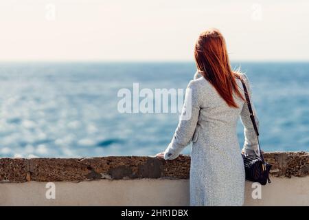 Die rothaarige Frau in Winterkleidung blickt auf das Meer Stockfoto