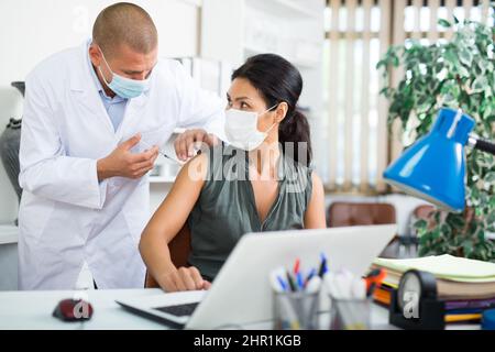 Arzt impft asiatische Geschäftsfrau am Schreibtisch im Büro Stockfoto