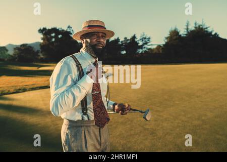 Ein Porträt eines eleganten, reifen schwarzen Mannes in elegantem Outfit mit einem weißen Hemd, einer Krawatte und einer Hose mit Hosenträgern, der auf einem abendlichen Golffeld steht Stockfoto