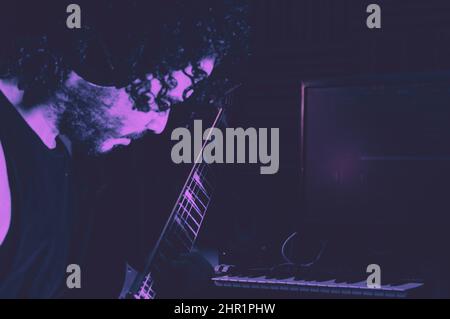 Seitenansicht des kaukasischen Mannes im Dunkeln, der E-Gitarre spielt, mit violetter Beleuchtung, mit Tastatur und Computer im Hintergrund, Kopierraum. Stockfoto
