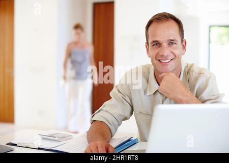 Arbeiten rund um die Uhr. Aufnahme eines reifen Mannes, der von zu Hause aus mit seiner Frau im Hintergrund arbeitet. Stockfoto