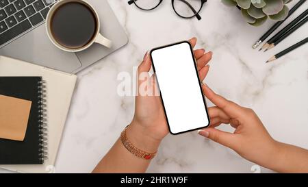 Eine Frau hält Smartphone leer Display Mockup für Montage Ihrer Grafik auf dem Bildschirm über ihrem weißen Marmor Schreibtisch. Draufsicht Stockfoto