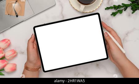 Weibliche Hände zeigen Tablet weißen Bildschirm mockup über ihrem schönen Schreibtisch. Laptop, Kaffeetasse, Gläser, Tulpenblüten und Einrichtung aus weißem Marmor b Stockfoto