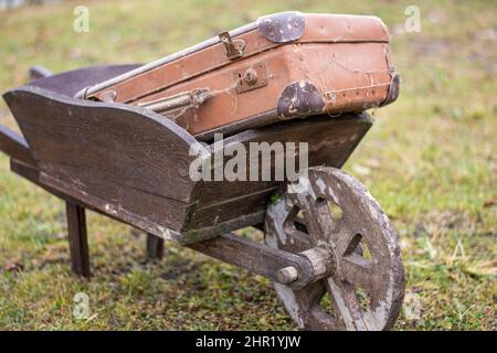 Brauner alter klassischer Retro-Vintage-Koffer auf einer hölzernen Schubkarre im Garten. Stockfoto