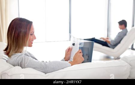 Die neueste Mode. Eine junge Frau liest ein Magazin, während ihr Freund im Hintergrund einen Laptop durchstöbert. Stockfoto