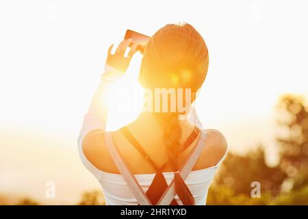 Ich bin froh, dass ich diese tolle Aussicht einfangen kann. Rückansicht einer unkenntlichen Frau, die in der Natur mit ihrem Handy ein Foto gemacht hat. Stockfoto