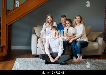 Große glückliche Familie. Großeltern im Porträt, Vater und ihre beiden lieben Kinder, Schwester und Bruder, sitzen zusammen auf der Couch zu Hause im Wohnzimmer und lächeln für die Kamera. Hypothekendarlehenkonzept Stockfoto