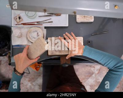 Eine Erwachsene Frau, die in ihrer handwerklichen Schmuckwerkstatt arbeitet, sitzt an einer Werkbank und verwendet Steinblöcke für ihre Arbeit. Stockfoto