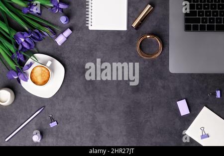 Frühling Frauen Arbeitsplatz Draufsicht mit schönen Irisblumen, Laptop, Bürobedarf, Beauty-Zubehör und Kaffee mit Milch auf dunkelgrauem Stein Hintergrund. Flach liegend mit Kopierplatz. Frühlingsferien, Muttertag und Geschäftskonzept. Stockfoto
