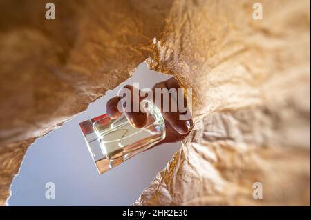 Eine Hand legt eine gelbe Flasche Parfüm in einen Papierbeutel. Die Hand eines reifen Mannes hält die Glasflasche über dem offenen braunen Beutel. Von unten nach oben geschossen. Stockfoto