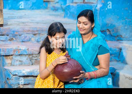 Glückliche indische Tochter legte Münze Ton Geldkasten oder gullak, während ihre Mutter in der Hand hielt. Lächelnde Frau trägt Sari Mädchen mit traditionellen Sparschwein, Stockfoto
