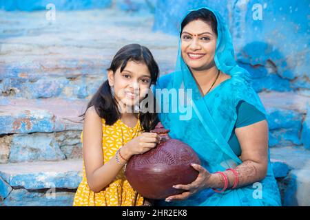 Porträt einer glücklichen indischen Mutter und Tochter, die Tongeldkiste oder Gullak in der Hand hält. Lächelnde Frau trägt Sari und Mädchen legte Münze in traditionelle Schwein Stockfoto