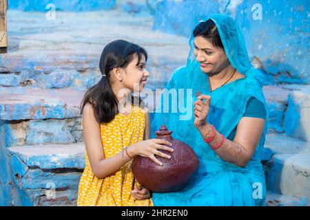 Glückliche indische Frau, die eine Münze hält, um sie in einen Tongeldkasten oder Gullak zu stecken, während ihre Tochter mit ihr sitzt. Lächelnde Frau trägt Sari mit traditionellem p Stockfoto