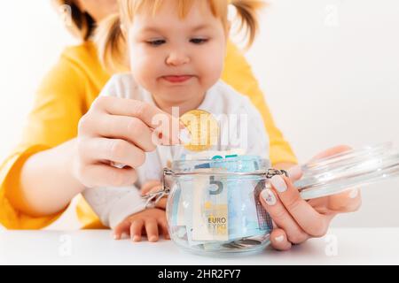 Das kleine Mädchen und ihre Mutter setzen Bitcoin in ein Glas mit Währung. Nahaufnahme. Das Konzept des Investierens und Online-Banking. Stockfoto