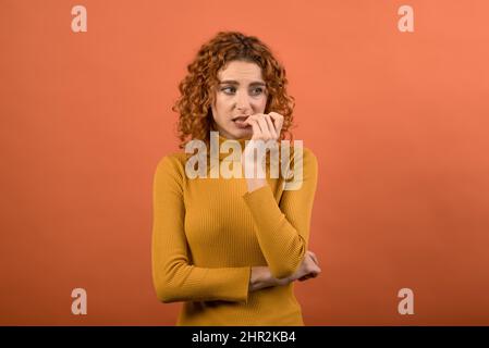 Ein junges und attraktives, rothaariges kaukasisches Mädchen in einem orangefarbenen Pullover, das ihre Fingernägel auf die Hand beißt, hat Angst, isoliert auf einem orangefarbenen Studiorücken Stockfoto