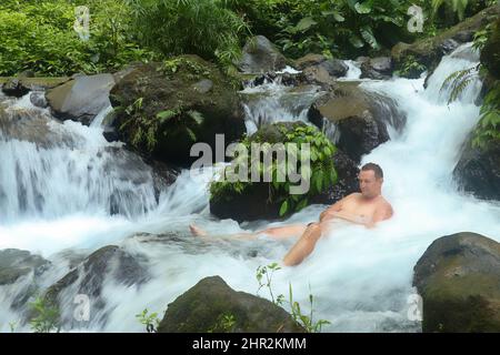 Ein junger Mann spritzt Wasser in den Fluss. Ein lächelnder junger Mann badet im Flusslauf Stockfoto