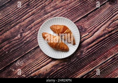 Ein paar Croissants auf einem weißen, strukturierten runden Teller auf einem Holzhintergrund von oben, ein Favorit beim Frühstück weltweit. Stockfoto
