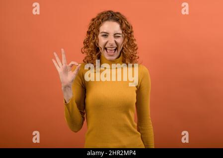 Junge und attraktive rothaarige kaukasische Mädchen in orange Jumper zeigt Finger Okay Geste isoliert auf Studio orange Hintergrund. Stockfoto