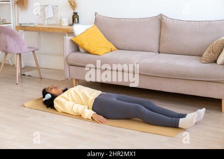 afroamerikanische Frau, die im Wohnzimmer auf einer Matte liegt. Entspannen Sie sich nach dem Training mit Kopfhörern Stockfoto