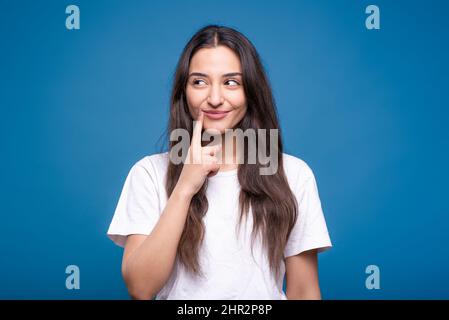 Attraktive kaukasische oder arabische Brünette Mädchen in einem weißen T-Shirt halten einen Finger in der Nähe ihrer Lippen, denken und lächeln isoliert auf einem blauen Studio Backgroun Stockfoto