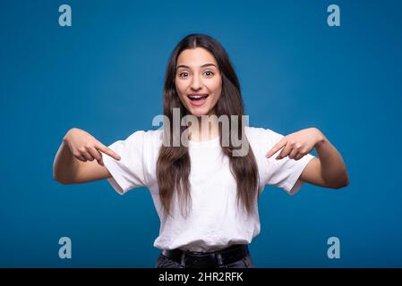 Junge und attraktive kaukasische oder arabische Brünette Mädchen in weißem T-Shirt zeigt die Finger nach unten auf Kopierfläche für Werbung isoliert auf blauem Studio Rücken Stockfoto