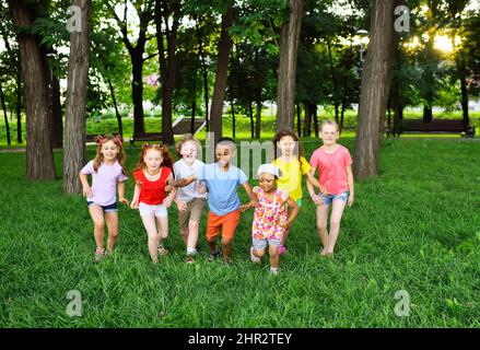 Eine Gruppe von Kindern von Jungen und Mädchen verschiedener Rassen laufen auf dem grünen Gras vor dem Hintergrund eines Erholungsparks. Kindertag, Stockfoto