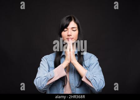 Attraktive kaukasische Brünette Mädchen in einem Hemd in einer meditativen Position stehend und zeigt ihre Hände in einer Gebetsture isoliert auf einem schwarzen Studio b Stockfoto