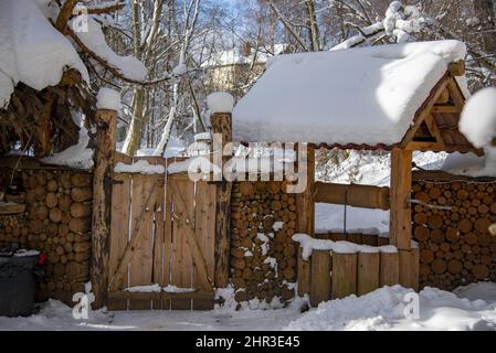 Ein Holztor in einem Blockzaun und ein Brunnen auf dem Hintergrund schneebedeckter Bäume. Stockfoto