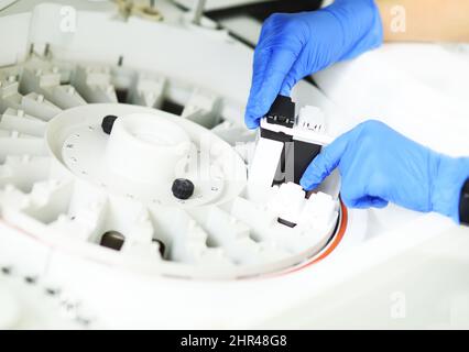 Laborassistent in blauen Gummihandschuhen fügt der Zentrifuge eines modernen biochemischen Analysators ein Reagenz hinzu. Stockfoto