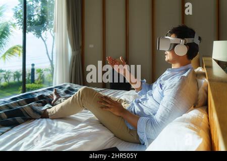 Junger asiatischer Mann, der während seines Sommerurlaubs auf dem Bett liegt und eine Virtual-Reality-Brille für Geschäftstreffen verwendet. Stockfoto