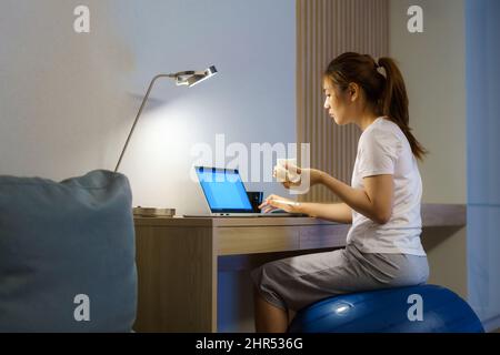 Asiatische Frau, die sich beim Yoga-Ball entspannt und Sandwich isst und sich mit Kollegen auf dem Laptop online unterhält, während sie nachts von zu Hause aus arbeitet. Stockfoto