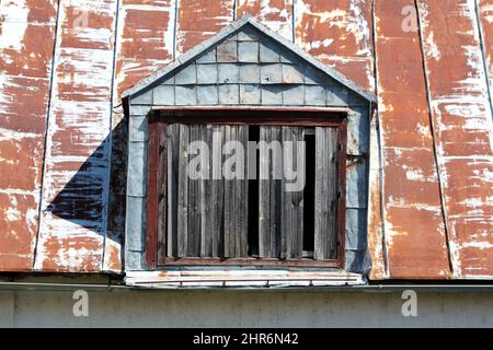 Vertäfelte beschädigte alte Dachfenster mit verfallenen dunkelbraunen gesprungenen Holzfensterrahmen, der mit einem kleinen Blechschutz und großen Rosten bedeckt ist Stockfoto