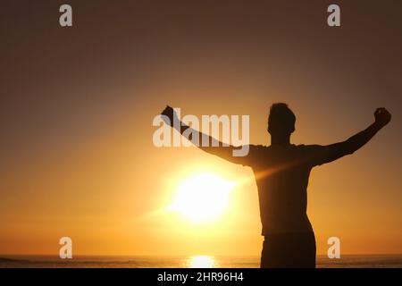 Sich von der Furche befreien. Silhouette eines Mannes mit ausgestreckten Armen und dem Sonnenuntergang vor ihm. Stockfoto