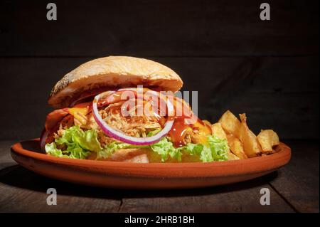 Pulled Pork Burger mit Salat, Käse, Zwiebeln und Barbecue-Sauce, serviert auf einem rustikalen Teller auf einem Holztisch. Hochwertige Fotografie. Stockfoto