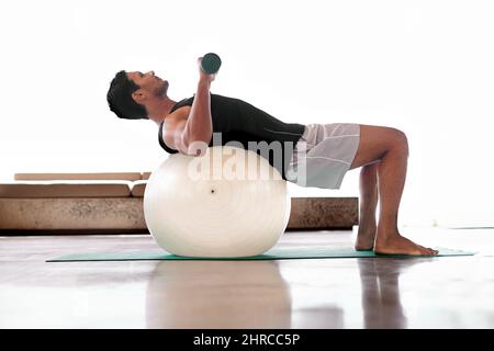 Er fand einen Weg, das Liegen zu einem Workout zu machen. Ganzkörperaufnahme eines Mannes, der Gewichte hebt, während er auf einem Übungsball liegt. Stockfoto