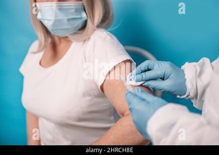 Der Arzt macht eine Impfung gegen ältere Frauen, die Gesichtsmaske auf blauem Hintergrund trägt. Impfung älterer Menschen. Stockfoto