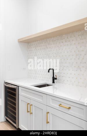 Eine Wet-Bar mit schwarzem Wasserhahn, weiß-goldener Mosaikfliese, weißen Schränken und einem Weinkühlschrank aus Edelstahl. Stockfoto