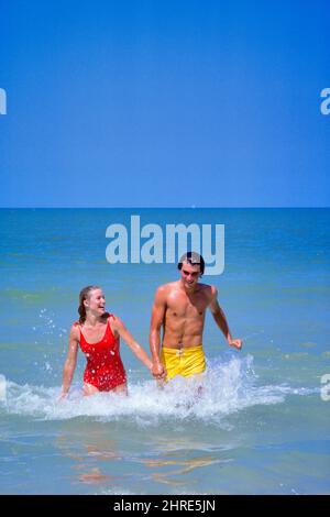 1980S LÄCHELNDES NASSES PAAR, DAS DIE HÄNDE HÄLT, DIE AUS DEM MEERWASSER GEHEN FRAU IN ROTEM BADEANZUG MANN IN GELBEM BAD BADEHOSE - KB15787 PHT001 HARS BADEN JUNGER ERWACHSENER TEAMARBEIT URLAUB FREUDE LIFESTYLE MEER WEIBCHEN SCHWIMMEN GESUNDHEIT KOPIEREN RAUM FREUNDSCHAFT HALBLANGE DAMEN PERSONEN MÄNNER SOMMERZEIT ZEIT OFF SHORE TRÄUME GLÜCK FRÖHLICH HOCHWINKEL ABENTEUER FREIZEIT REISE AUFREGUNG ERHOLUNG STRÄNDE URLAUB BEZIEHUNGEN LÄCHELT SCHWIMMER VERBINDUNG BEWEGUNG UNSCHÄRFE KONZEPTUELLE FLUCHT FREUDIG SANDIG STILVOLLE FLITTERWOCHEN BRAUTPAAR SAISON ZWEISAMKEIT KOFFER URLAUB JUNG ERWACHSENER MANN Stockfoto