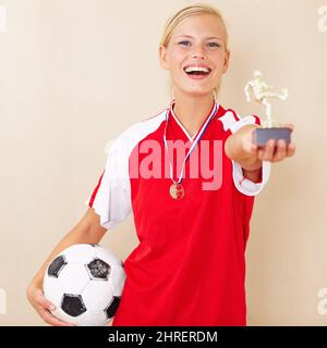 Gewinnen kommt ihr leicht. Porträt einer jungen Frau in einer Fußballuniform mit einer Trophäe und einem Fußball. Stockfoto