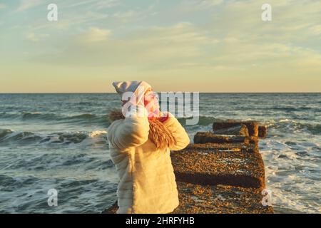Eine Frau mit blondem Haar in einem Hut und einem Pelzmantel geht an einem Steinbrecher entlang des Meeres Stockfoto