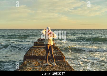 Eine Frau mit blondem Haar in einem Hut und einem Pelzmantel geht an einem Steinbrecher entlang des Meeres Stockfoto
