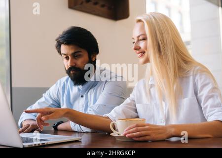 Seitenansicht einer lächelnden blonden Geschäftsfrau, die auf den Laptop-Bildschirm schaute und dem bärtigen indischen männlichen Kollegen die Punkte erklärte. Stockfoto
