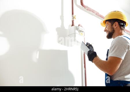 Blick in den niedrigen Winkel auf den Techniker, den Heizungsmonteur in Schutzhelm und Schutzhandschuhe, die Notizen machen, während der Gasverbrauch an Gasleitung und Messgerät überprüft wird Stockfoto