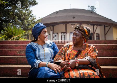 Zwei afrikanische Frauen unterhalten sich, sitzen auf der Treppe, beide sind in traditioneller Kleidung gekleidet Stockfoto