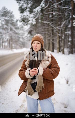 Junge stilvolle Millennial asiatische Frau in s Schaffell Mantel und Strickmütze auf Winterwald verschneiten Straße Stockfoto