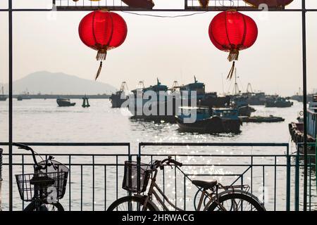 Traditionelle rote Laternen und Fahrräder umrahmen lokale Fischerboote am Ufer von Cheung Chau, einer abgelegenen Insel in den New Territories von Hongkong Stockfoto