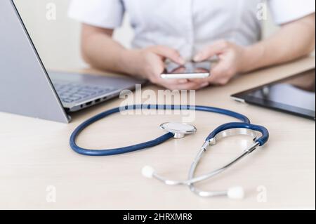 Nicht erkennbare Ärztin sitzt an einem Schreibtisch und nutzt ein Smartphone. Nahaufnahme der Hände eines Praktizierenden mit einem Telefon. Stethoskop, Tablet und Laptop Stockfoto