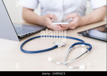 Nicht erkennbare Ärztin sitzt an einem Schreibtisch und nutzt ein Smartphone. Nahaufnahme der Hände eines Praktizierenden mit einem Telefon. Stethoskop, Tablet und Laptop Stockfoto