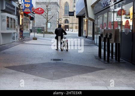 Stuttgart, Deutschland - 06. Januar 2022: Asiatischer Mann schob sein Fahrrad durch die Stadt. Straßenfoto mit Geschäften und Lebensmittelgeschäften am frühen Morgen Stockfoto